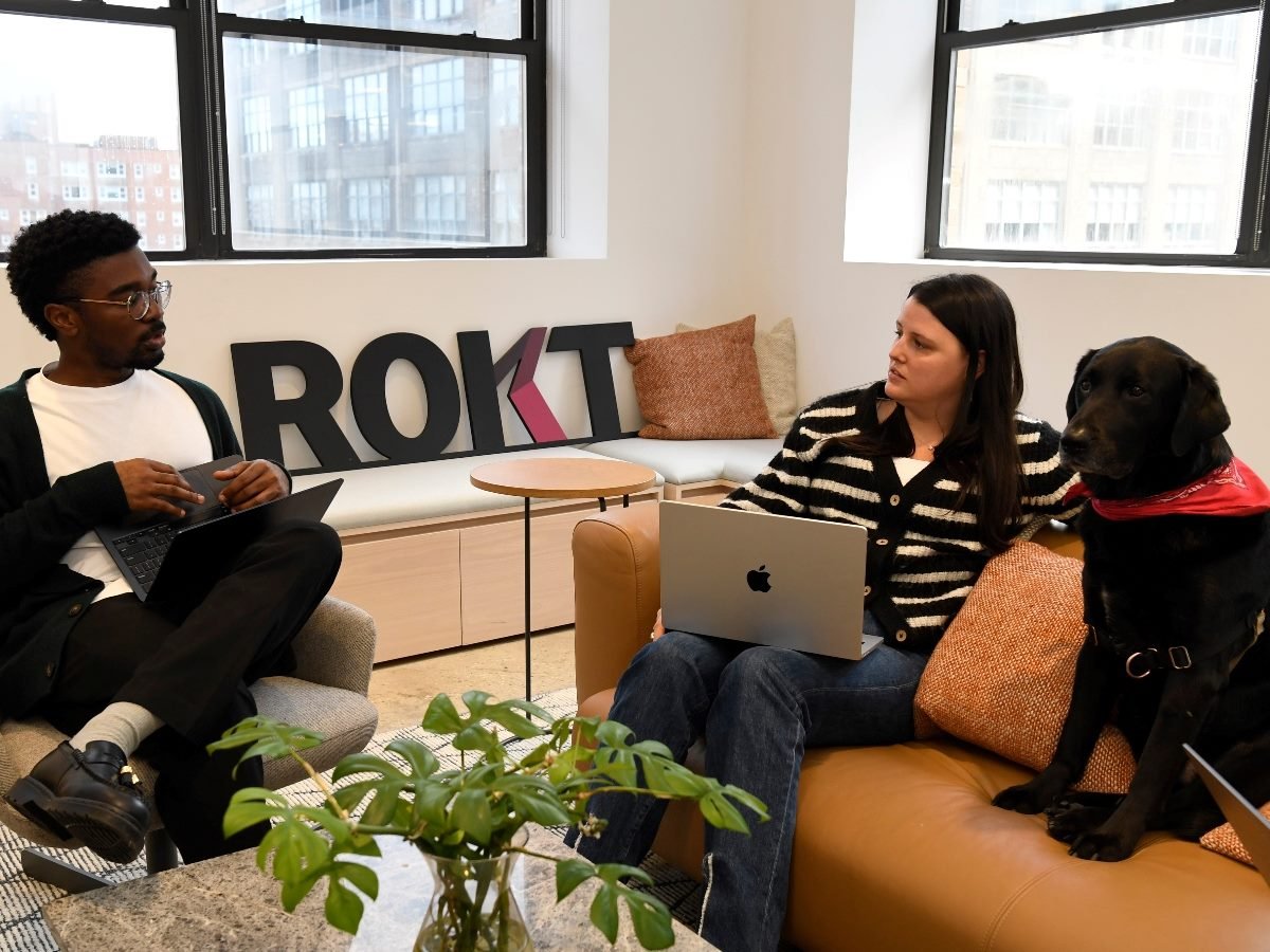 Two Rokt employees sit in a modern office with laptops, engaged in discussion beside a black dog wearing a red bandana. The Rokt logo is displayed in the background.
