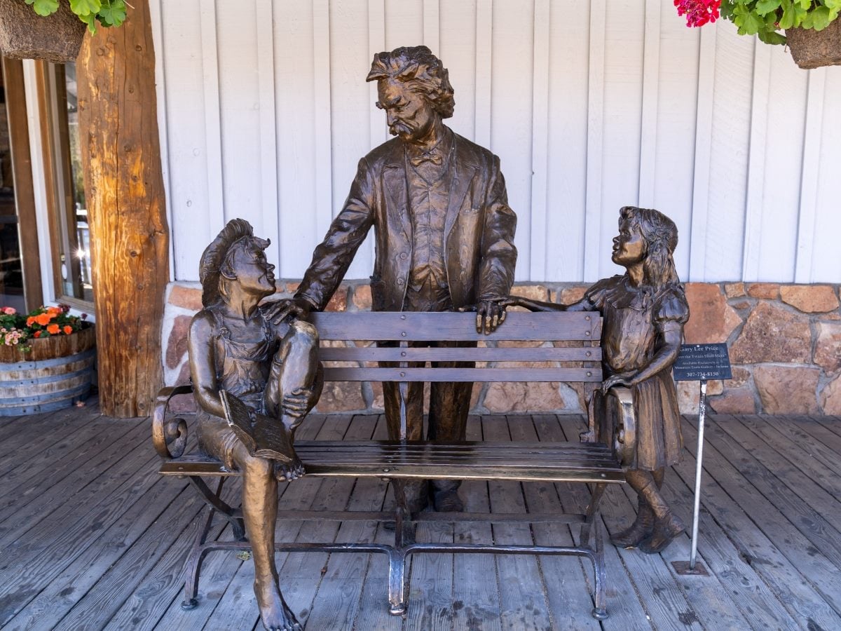 A bronze sculpture of Mark Twain standing behind a park bench, resting one hand on the shoulder of a barefoot boy seated on the bench reading a book and the other hand near a young girl standing beside the bench, all cast in dark bronze patina; the scene sits on a wooden porch deck against a white board-and-batten storefront wall with a stone foundation, flanked by hanging flower baskets and potted flowers, with a small placard identifying the artwork mounted on a stand near the bench.