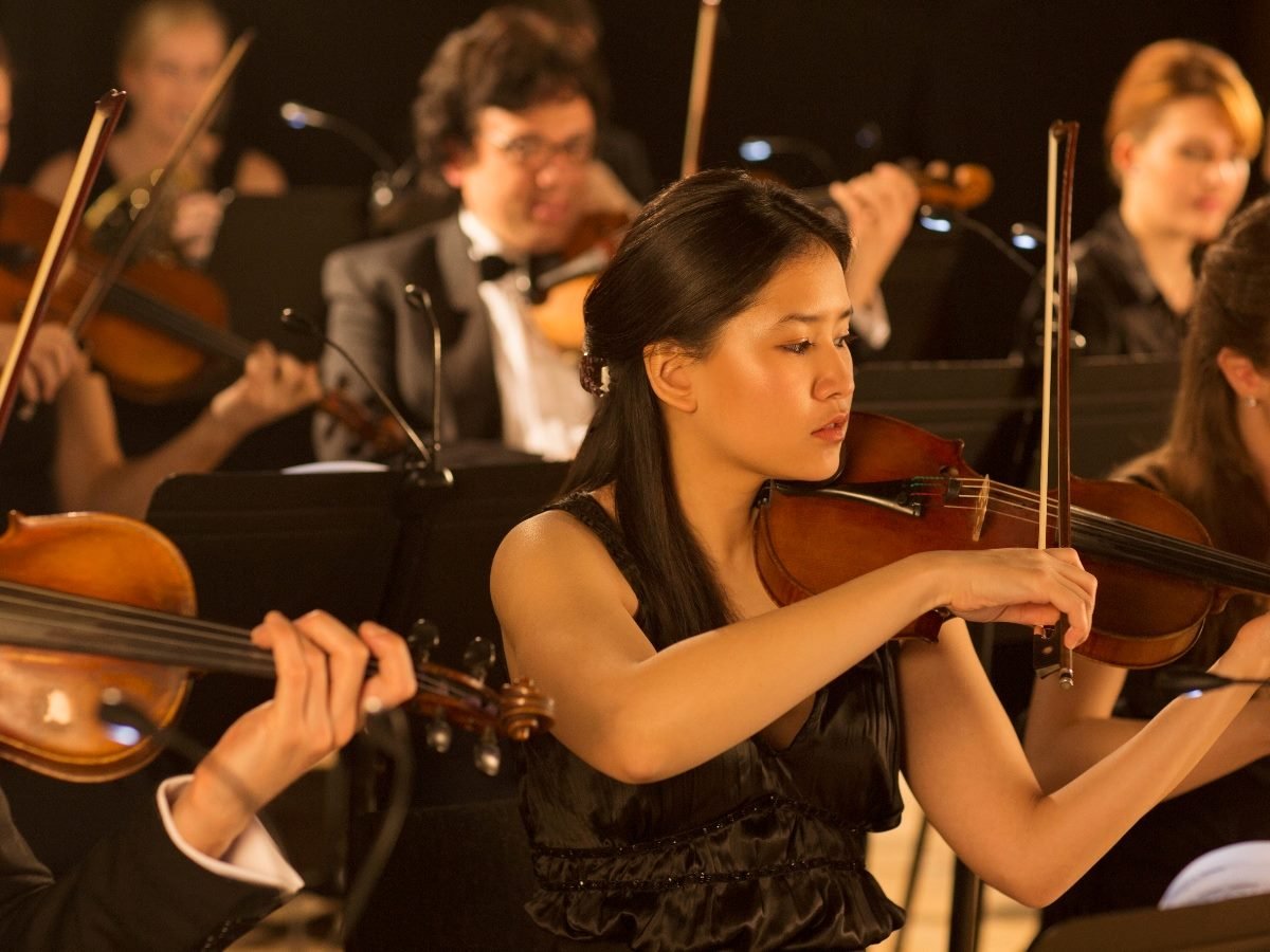 A focused violinist plays in an orchestra, surrounded by fellow musicians performing string instruments during a concert.