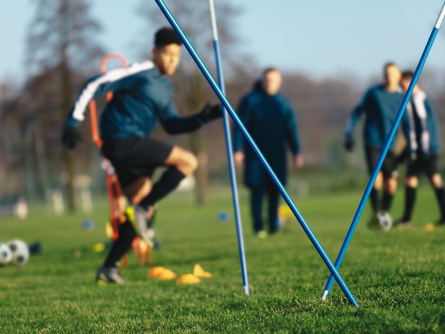 Soccer training session for junior level athletes with agility sticks and a player trying to maneuver in and around them. Other players in background on grass turf. 