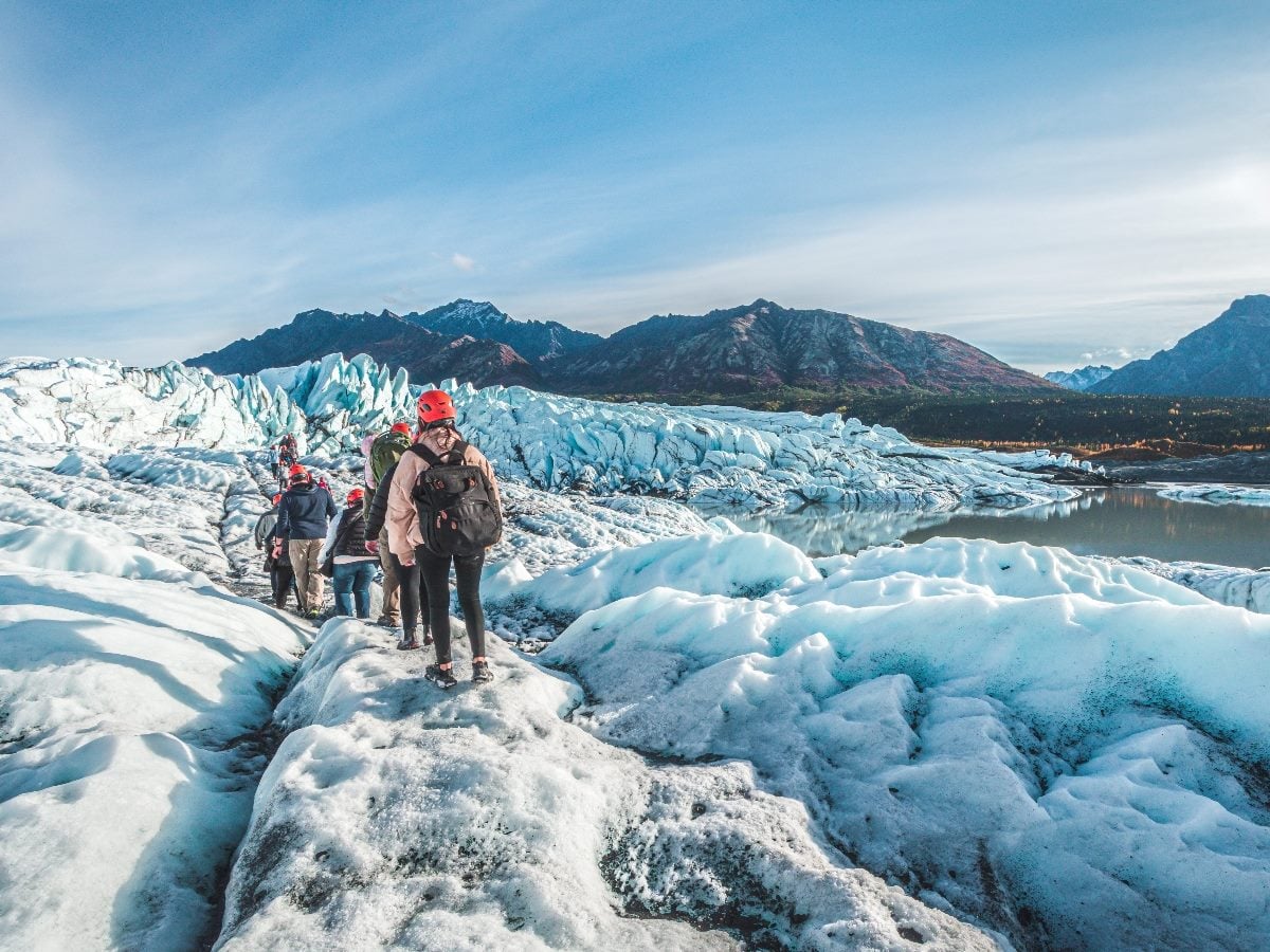 Guided hikers wearing helmets and backpacks walk single file across the icy surface of Matanuska Glacier in Alaska, with rugged mountains and a glacial lake in the background under a clear blue sky.