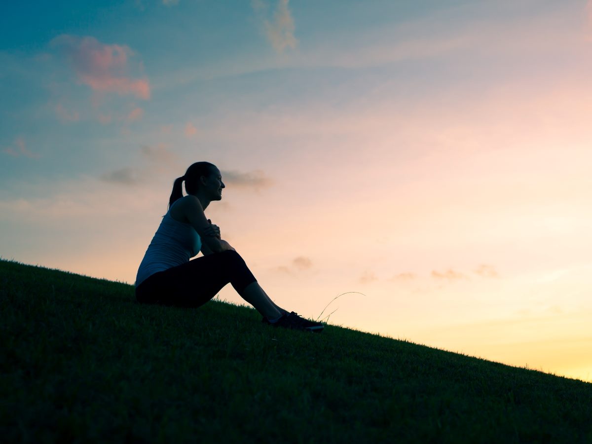 Silhouette of a woman sitting thoughtfully on a grassy hill at sunset, with a calm sky in the background.