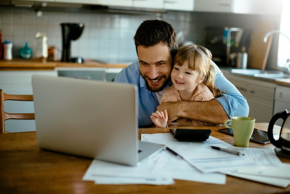 A young father embraces his child while sitting in front of a laptop computer at a kitchen table with coffee cups, pens and papers littering the table top in piece about trust in AI.