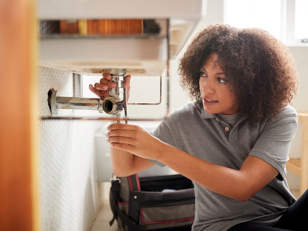 A woman crouches under a kitchen sink, using a wrench to tighten exposed plumbing pipes, with a toolbox beside her as she focuses on the repair.