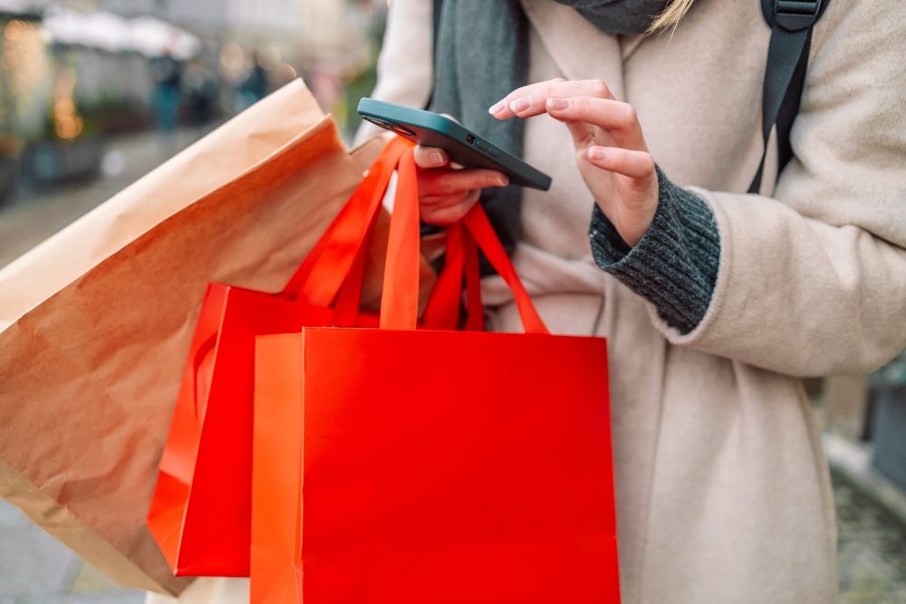 Cropped image of a person carrying three bags, one tan and two red, and typing on a smartphone in piece about user experience analysis and customer journeys.