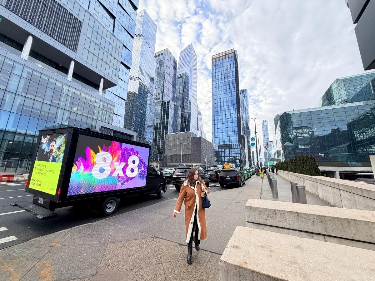 A digital advertising truck displaying colorful 8x8 branding drives through a busy city street lined with glass office towers, while a pedestrian in a brown coat walks along the sidewalk in the foreground.