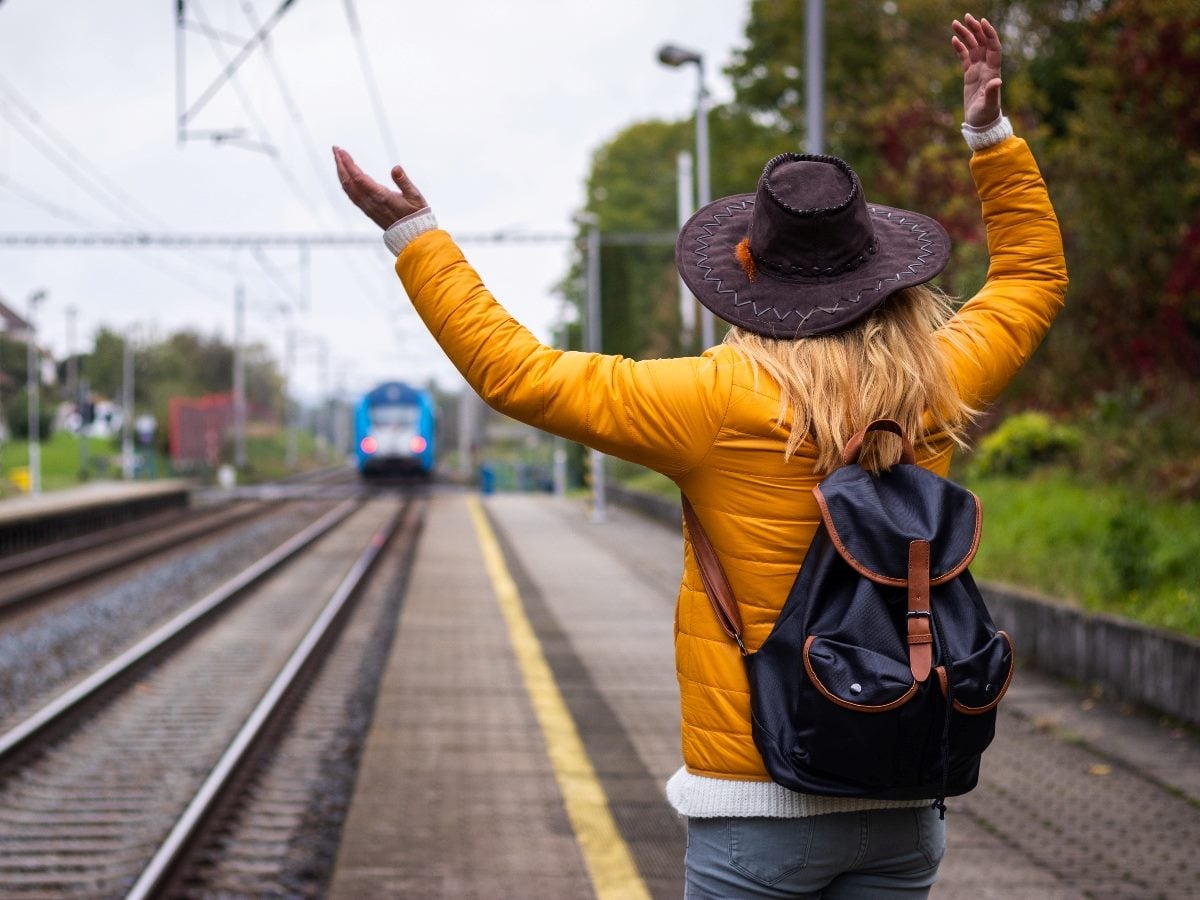 Person wearing a yellow jacket, backpack and wide-brimmed hat stands on a train platform with both arms raised as a departing train recedes into the distance, illustrating a missed connection or missed opportunity.