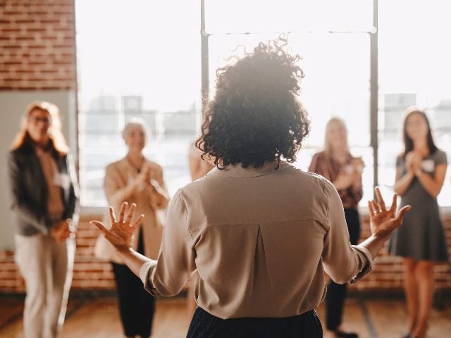 Shot from behind a woman leading a support-group class with five other woman blurred in background.