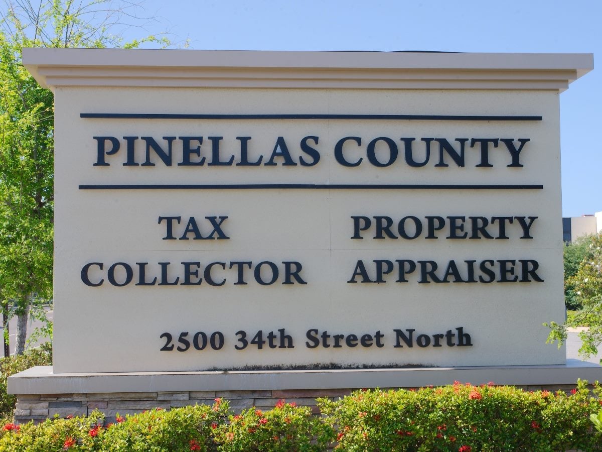 A large outdoor sign for Pinellas County displaying “Tax Collector” and “Property Appraiser” with the address 2500 34th Street North, surrounded by landscaping and trees under a clear blue sky.