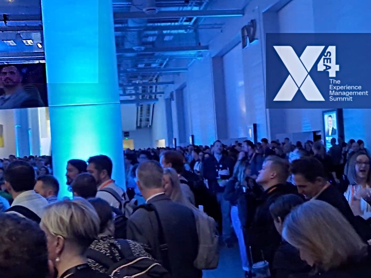 A large crowd gathers inside a blue-lit conference hall at the Qualtrics X4 Experience Management Summit at the Seattle Convention Center March 18, 2026, with a prominent X4 logo displayed on a screen, emphasizing the scale and energy of the event.