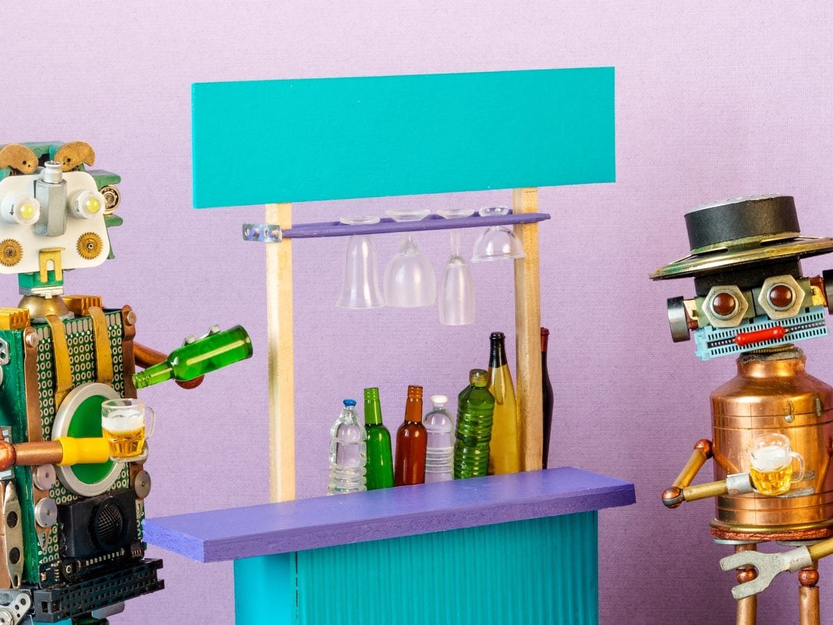 Two whimsical robot figures appear to socialize at a miniature bar setup, each holding a small beer mug. One robot pours from a green bottle while another wears a hat and faces the counter. The colorful scene features toy bottles, hanging glasses and a playful handcrafted design against a light purple background.