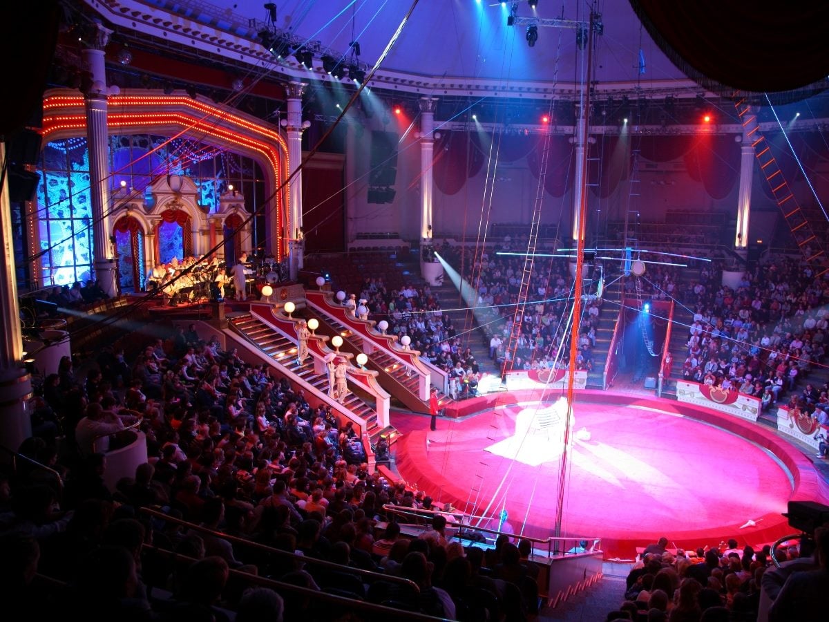 Wide view inside a packed circus arena with a bright red center ring, overhead rigging, dramatic blue and red stage lighting, ornate balconies, and a live band performing beside the ring.