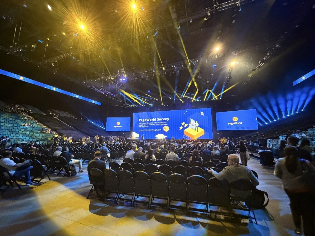 Wide-angle view inside the PegaWorld keynote arena before a main session begins, showing attendees taking seats in a large auditorium. Blue PegaWorld-branded screens dominate the stage while yellow and blue spotlights illuminate the venue from overhead rigging. Rows of black chairs fill the floor as conference participants gather and converse ahead of the presentation.