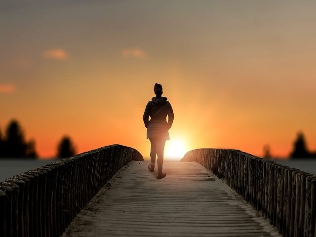 A girl stands on bridge lake sunset; shot taken behind the girl. 