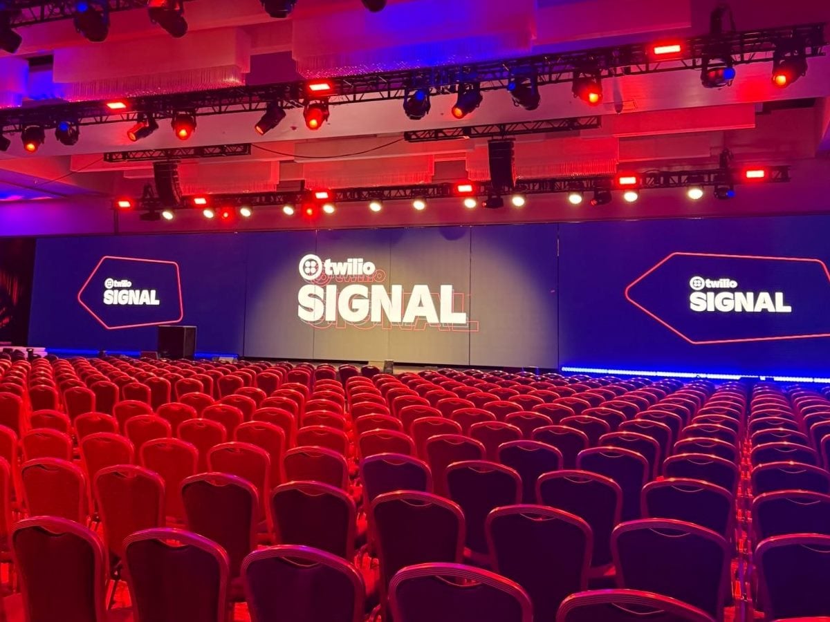 ALT TEXT The main stage at Twilio SIGNAL 2026 in San Francisco, with rows of empty red chairs facing a large center screen displaying the Twilio SIGNAL logo, flanked by two matching branded side screens under red and blue stage lighting.