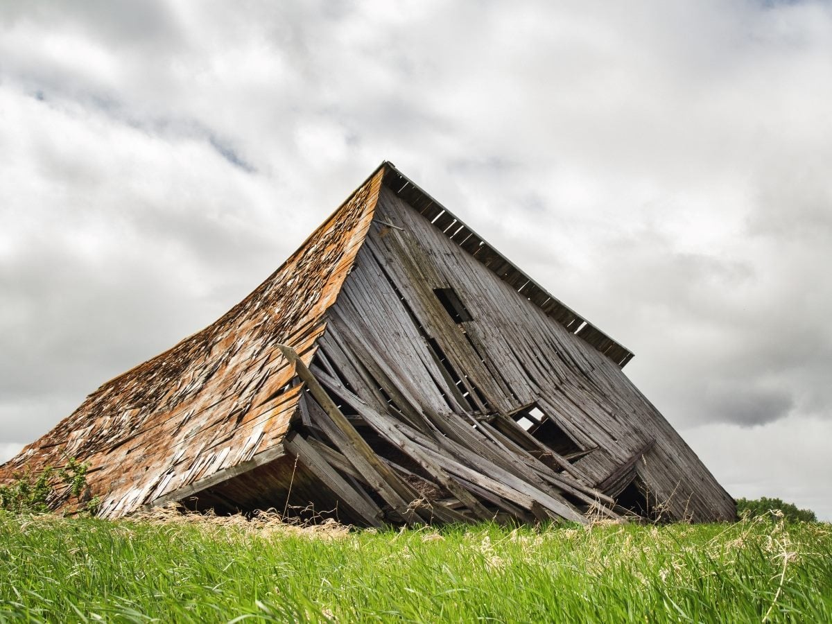 A weathered wooden barn with its roof and walls partially collapsed leans dramatically toward the ground in a grassy field. The aging structure shows broken boards, missing siding and open window gaps beneath a sky filled with thick gray clouds, illustrating severe structural deterioration and neglect.
