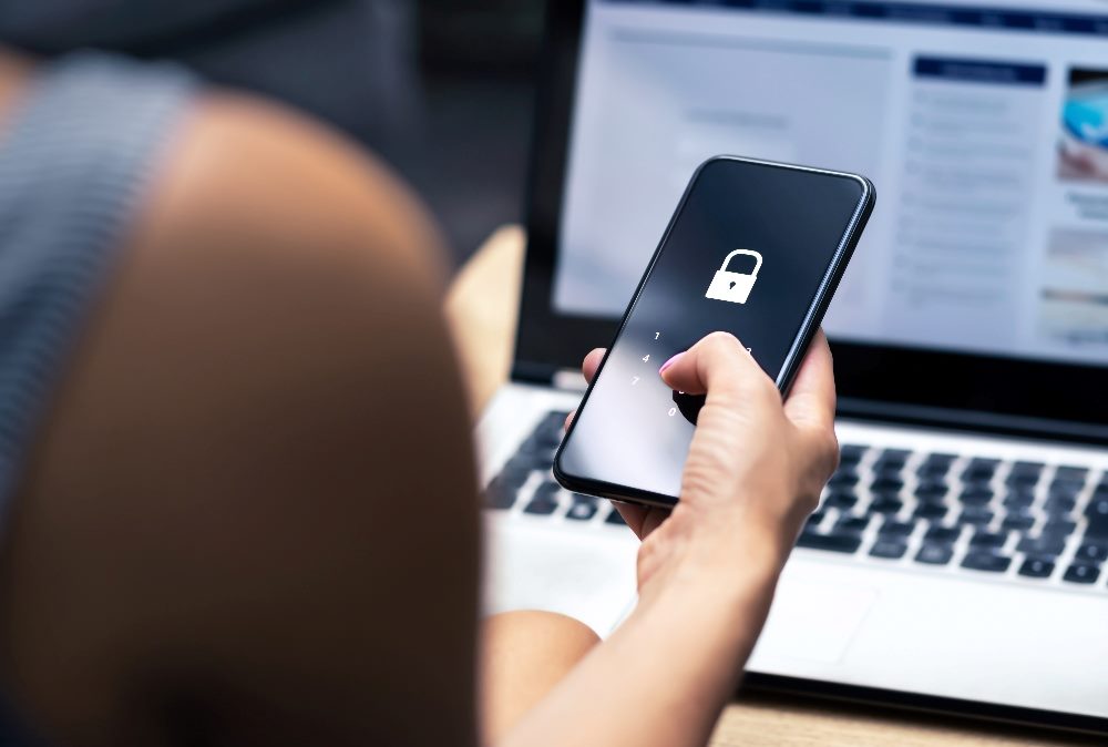 A person sits at a desk holding a smart phone with a lock symbol and keypad showing on the screen in their hand with a laptop in the background in a piece about third party cooke deprecation.