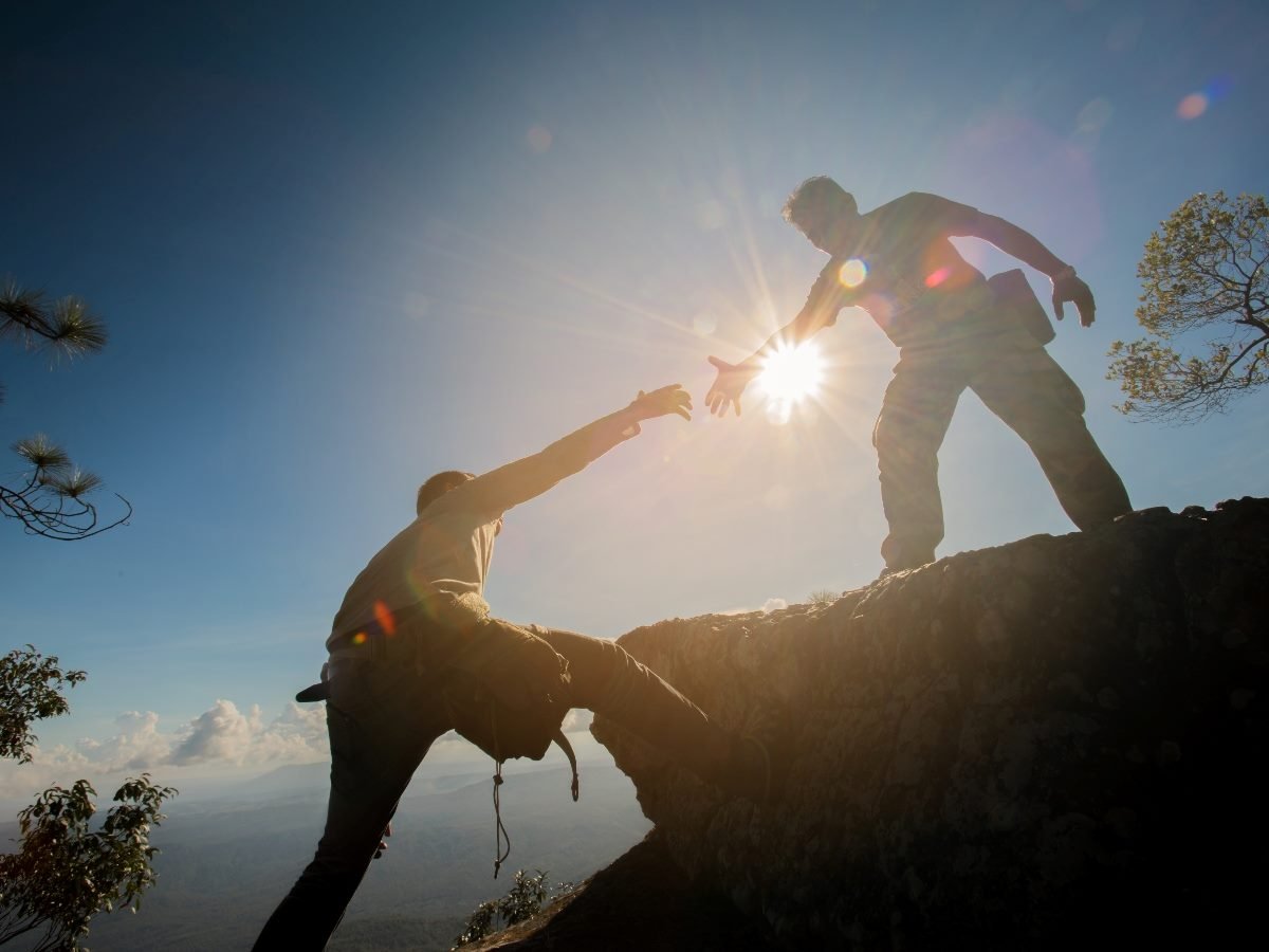 Two hikers silhouetted by the sun as one reaches down to help the other climb up a rocky ledge, symbolizing trust, support and partnership.