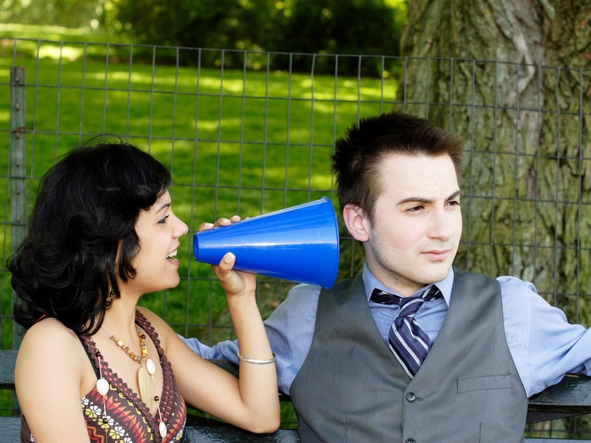 A woman speaks through a blue megaphone toward a man sitting beside her on a bench, while he looks away with disinterest, symbolizing ignored communication.