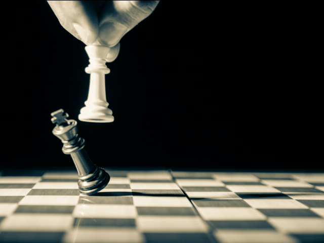 Black and white image of chessboard with white king taking down black king. Hand and fingers and chess board