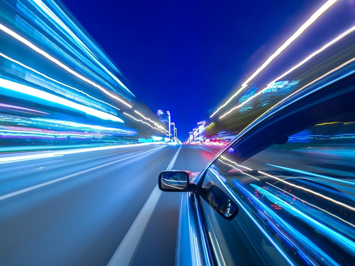 A high-speed nighttime driving scene viewed from the side of a moving car, with blue and white light trails streaking across the roadway and city lights blurred in the distance to convey speed, acceleration and motion.