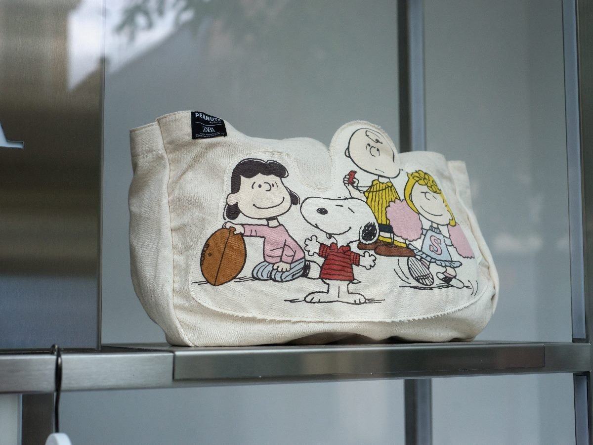 A canvas tote bag displayed on a shelf featuring Peanuts characters, including Snoopy, Lucy, Charlie Brown and Sally, printed on the front.