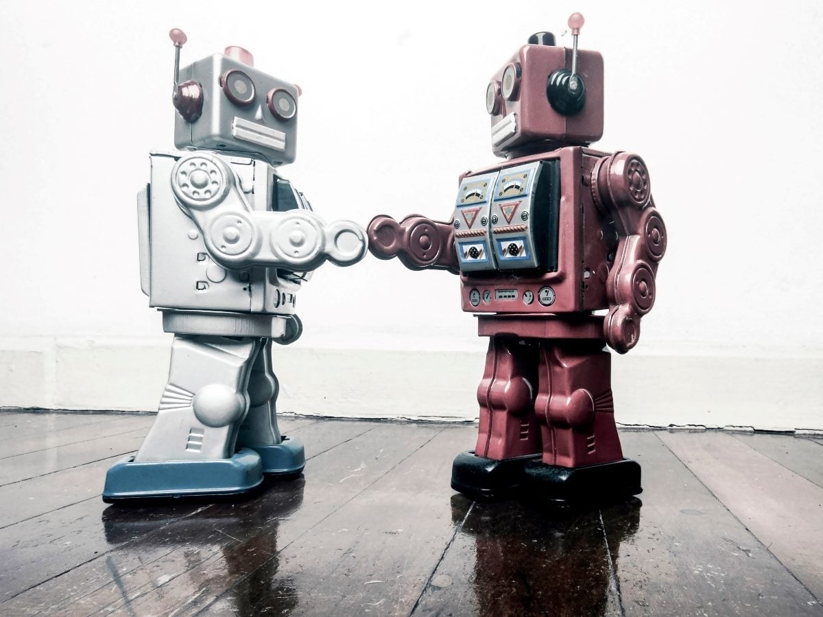 Two vintage toy robots face each other and shake hands on a polished wooden floor. One robot is silver with blue feet, while the other is maroon with decorative control panels on its chest. The plain white background keeps the focus on the retro mechanical figures and their gesture of cooperation.