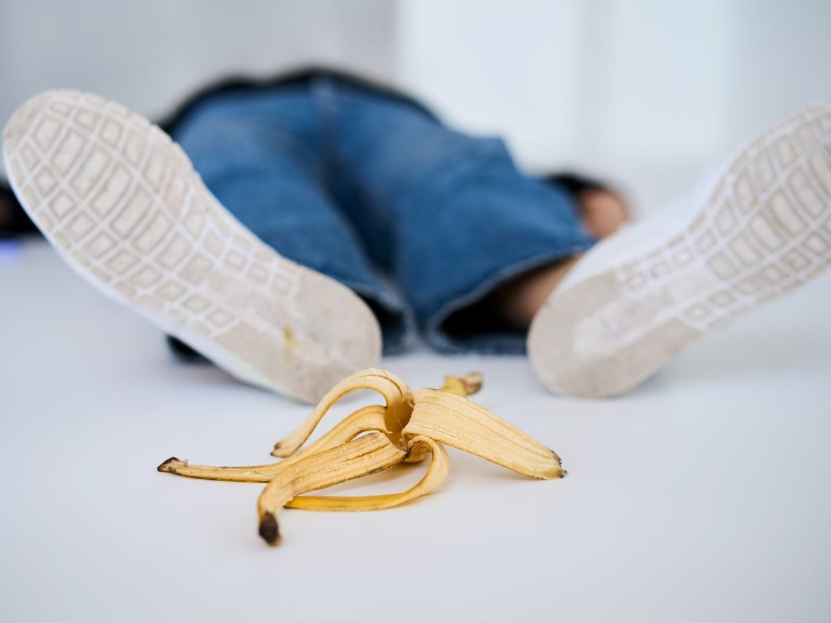 A person lying on the floor with legs extended, wearing jeans and white-soled shoes, after slipping on a banana peel in the foreground.