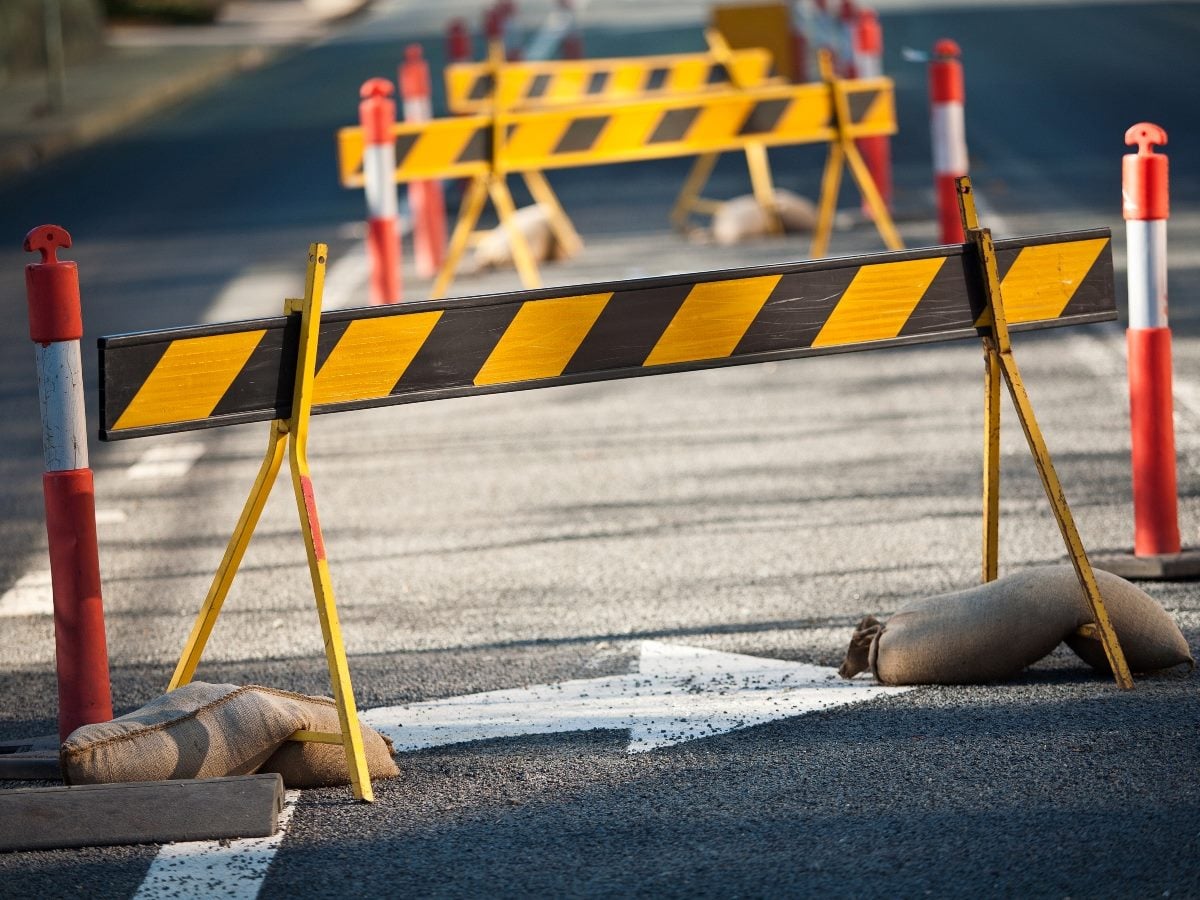 Two wooden A-frame barricades with black and yellow diagonal-striped crossbars block a paved road, set at a low angle so the nearer barricade fills most of the frame while a second, matching barricade sits blurred in the background. Red-and-white striped delivery posts flank both barricades on either side of the road. Tan sandbags weigh down the base of each barricade's legs, and a white directional arrow painted on the asphalt points toward the nearer barrier. The scene is lit by warm, low daylight casting long shadows across the road surface, with the background falling out of focus to keep attention on the foreground barricade.
