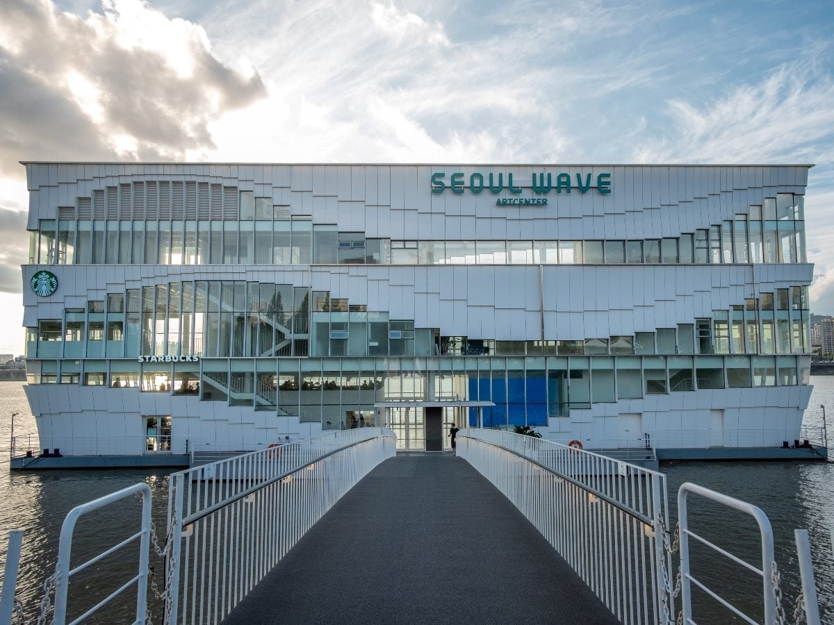 A daytime photograph of a large white floating building on a river, shot straight-on from a bridge leading up to its entrance. The structure's exterior is composed of angular, overlapping white paneling with a wave-like ripple pattern across the upper facade, and rows of glass windows span multiple levels. Green "SEOUL WAVE" and "ARTCENTER" signage sits at the top of the building, with a Starbucks logo and "STARBUCKS" wordmark visible on the lower left section behind glass storefronts. A long gray pedestrian walkway with white metal railings extends from the foreground toward a central glass entrance door, where a single person is visible walking toward the building. The structure floats on calm river water, flanked by white mooring platforms and life-ring safety equipment on either side. Above, a bright sky with scattered clouds and soft late-day sunlight fills the upper half of the frame, with distant city buildings faintly visible along the horizon on the right.