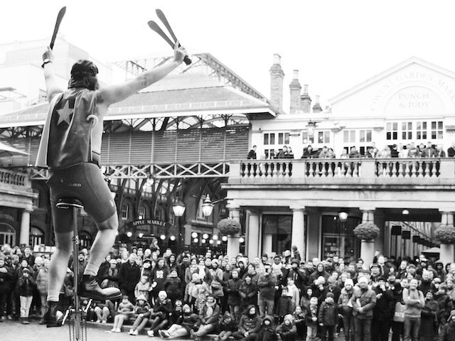 man on unicycle wearing a cape juggling swords in front of a crowd