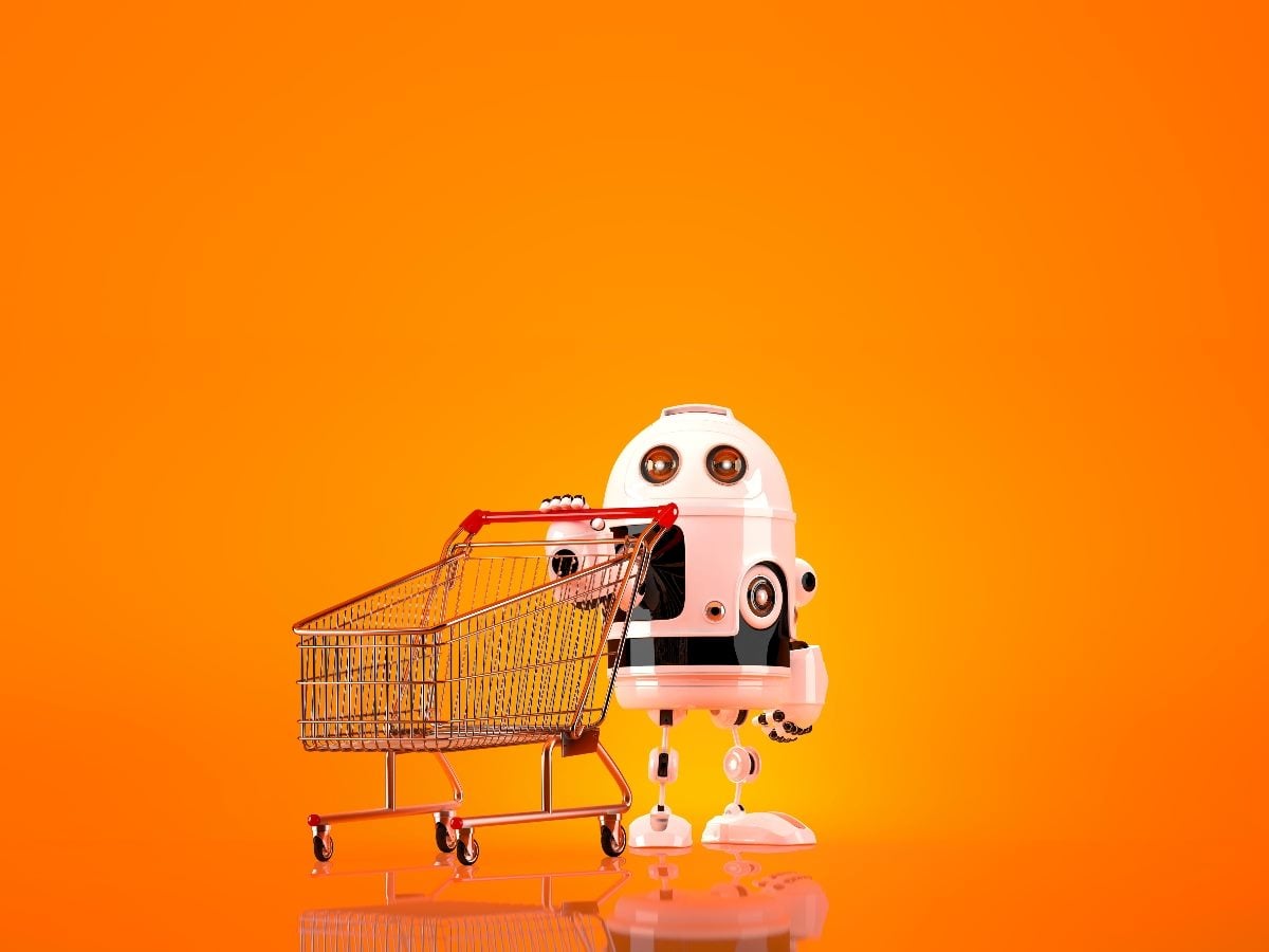 Small white robot with large round eyes stands beside an empty metal shopping cart against a bright orange background, symbolizing AI-powered shopping or agentic commerce.