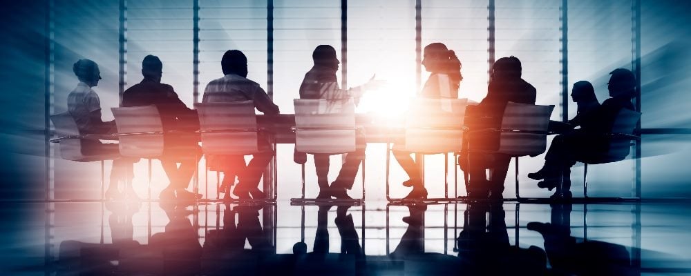 Businesspeople or stakeholders sit at a table for a meeting in a modern office with large glass windows and the sun setting in the background, suggesting the importance of marketing stakeholder meetings.