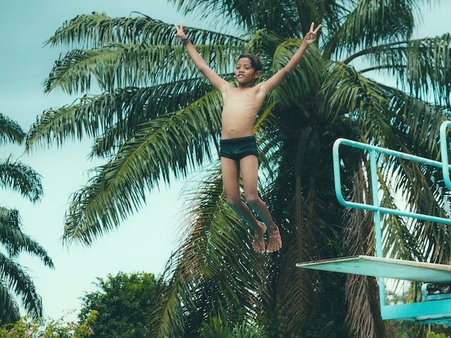 boy jumping off a diving board with hands in the air giving the peace symbol
