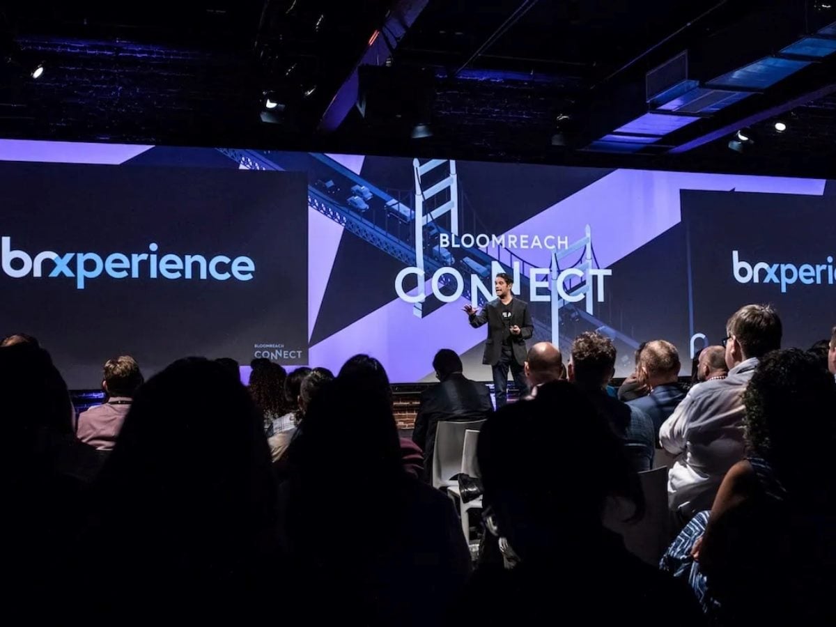 A speaker presents on stage at Bloomreach Connect, with large “brXperience” and “Bloomreach Connect” branding displayed on wide screens behind them, addressing an audience seated in a darkened conference venue.