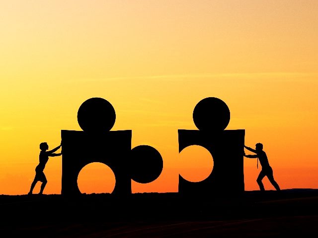 The silhouettes of two people push two giant puzzle pieces together against a dramatic sunset in piece about CX and EX synergy.