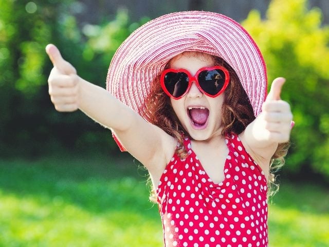 Young red-haired child smiling and wearing a red and white striped hat, a red and white polka dot shirt and red heart sunglasses giving two thumbs up in piece about marketing and CX goals and happy customers. 