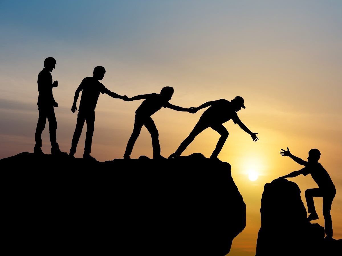 Silhouettes of several people standing on a cliff at sunset, reaching down to help pull another person up from a lower ledge, symbolizing collaboration, trust and support.