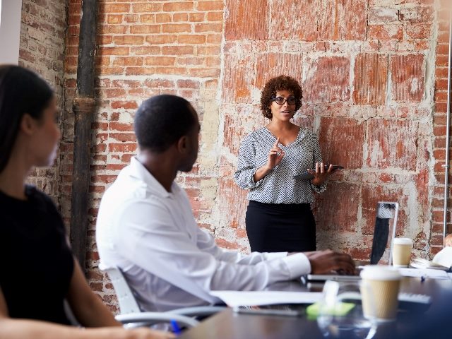 Business woman standing up leading a meeting in a conference room. Others sitting down at a conference table.