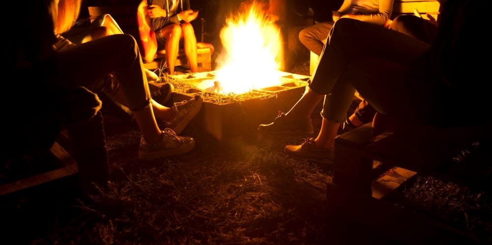Friends sit around the campfire talking and telling stories with orange fire burning in the center, representing the power of customer experience storytelling.
