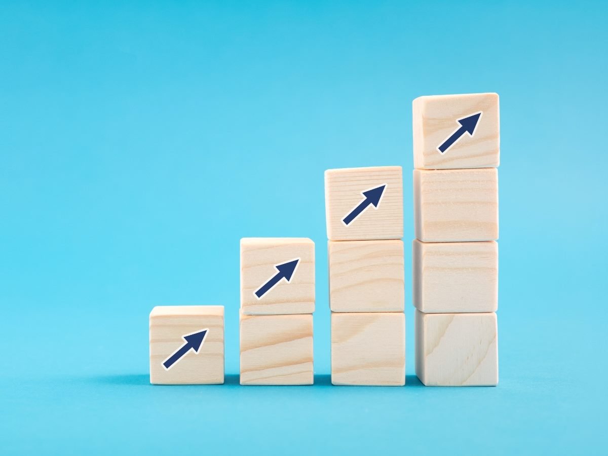 A row of wooden blocks arranged in ascending height against a blue background, each block displaying an upward-pointing arrow to represent business growth and progress.