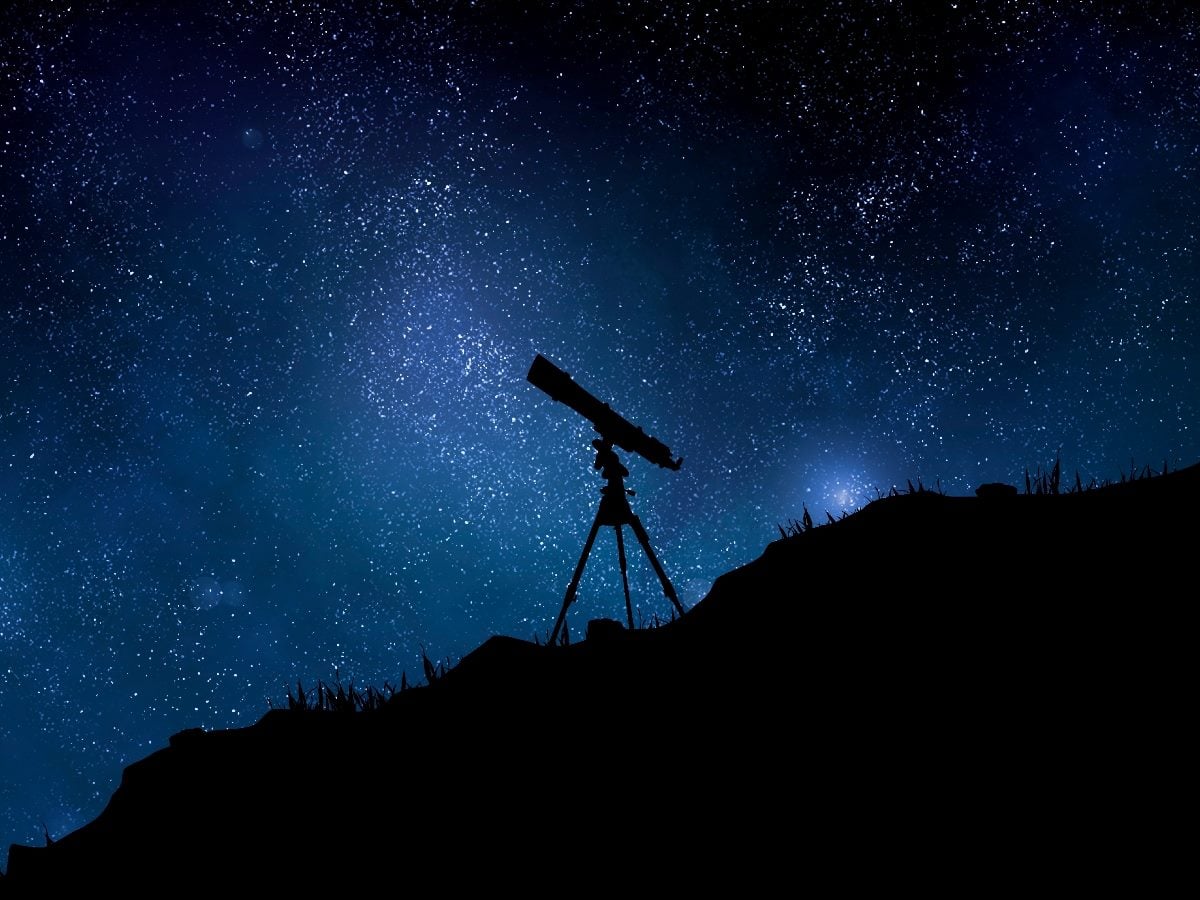 Silhouette of a telescope mounted on a tripod atop a hillside, set against a deep blue night sky filled with dense stars and the Milky Way galaxy.