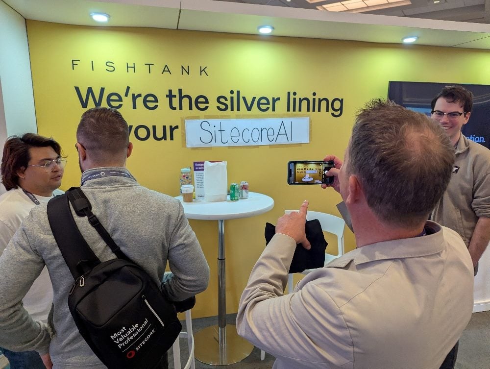 A group of conference attendees gathers at the Fishtank booth at Sitecore Symposium. A man takes a photo of a booth sign where “XM Cloud” has been humorously covered with a hand-written “SitecoreAI” label.
