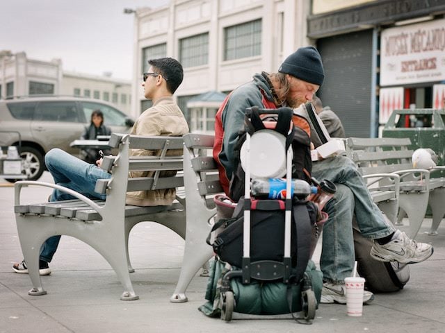 two men sitting back to back on benches who look to be living in very different circumstances