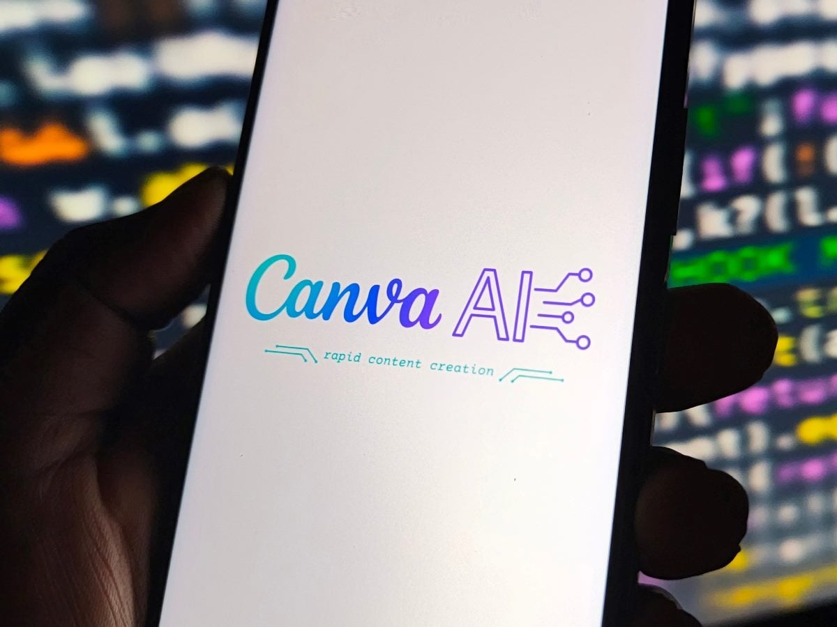 Close-up of a hand holding a smartphone displaying the Canva AI logo with the tagline “rapid content creation,” set against a blurred background of colorful digital screens.