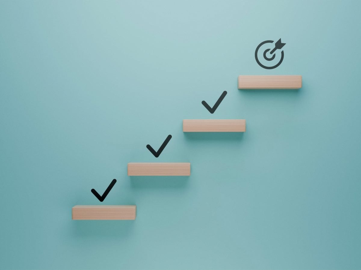 Four light wood plank steps ascend diagonally from lower left to upper right against a flat teal-blue background, each step topped with a dark checkmark icon except the highest, which instead holds a circular target icon with an arrow striking its center. The steps increase evenly in height, creating a rising staircase pattern with soft, even lighting and no visible shadows beyond subtle contact shading beneath each plank. The composition is shallow and graphic, with a shallow depth of field that keeps all elements in sharp focus against the softly lit, uncluttered background.