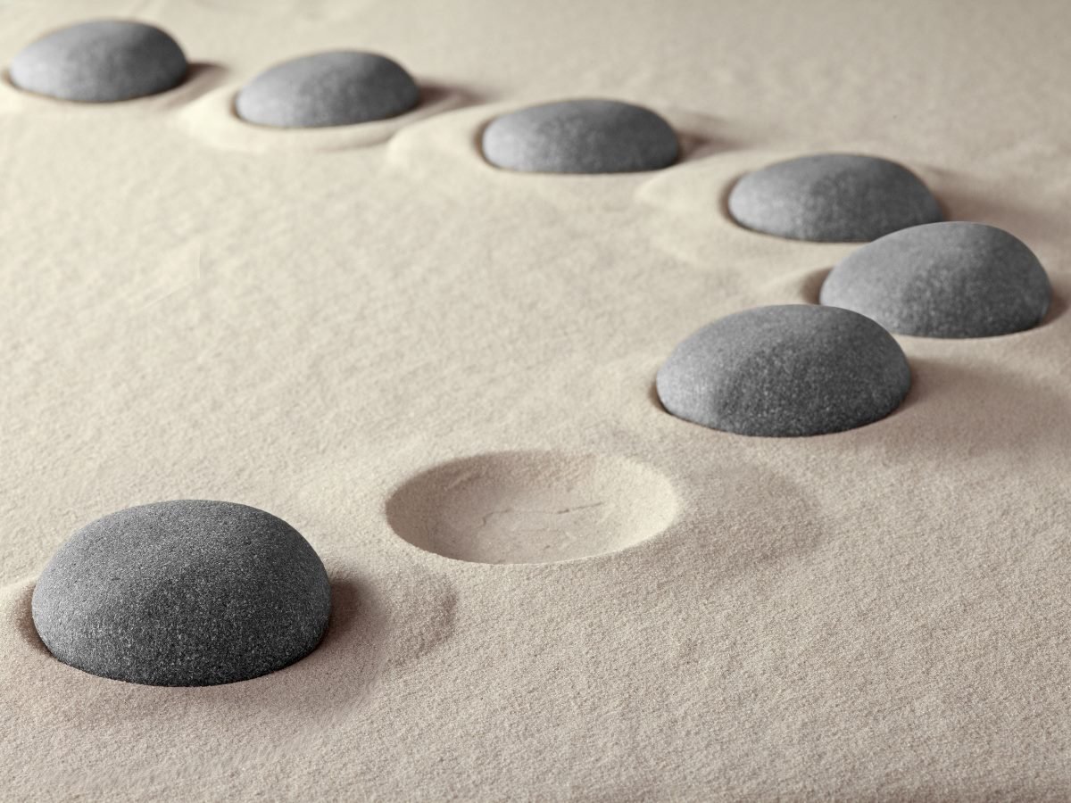 Smooth gray stones partially embedded in fine beige sand, arranged in a curved path with one empty indentation where a stone appears to be missing.