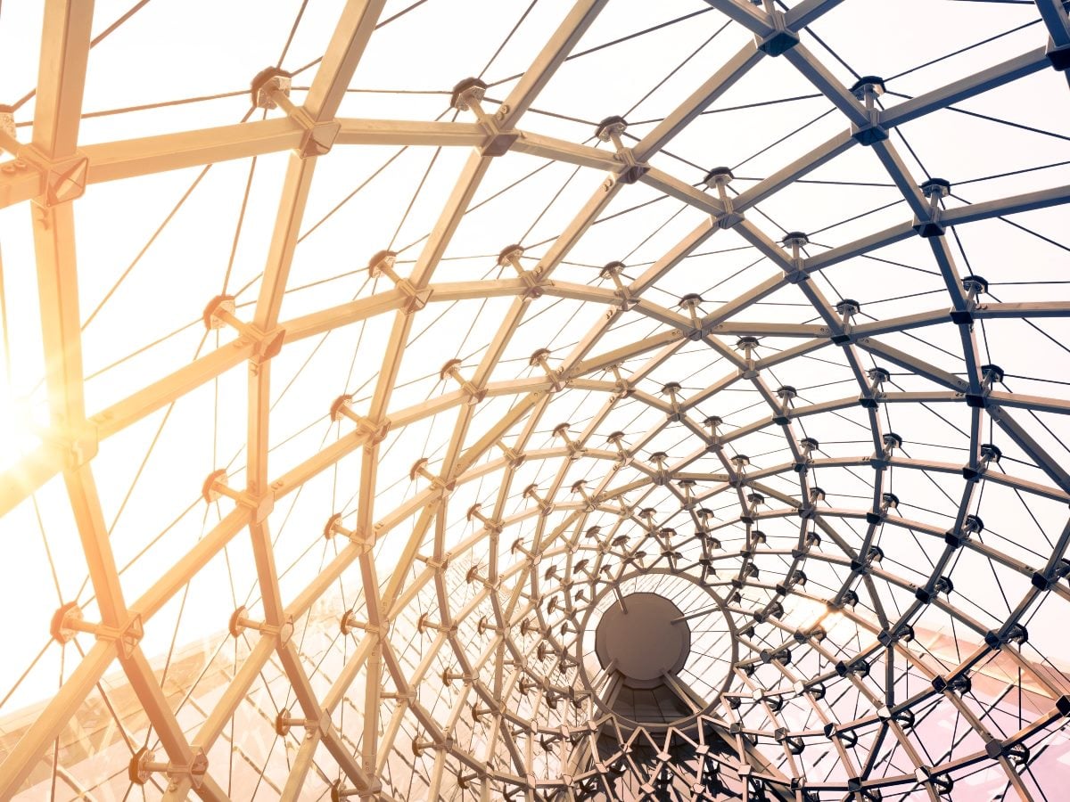 Abstract architectural image of a curved glass-and-steel framework with intersecting beams and cables forming a connected lattice. Sunlight streams through the structure, emphasizing its layered design and interconnected geometry, symbolizing the foundational framework and relationships required to support AI-ready marketing measurement.