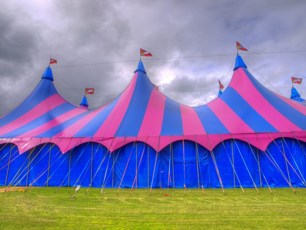 Huge big top red and blue circus tents surrounded by tent ropes and stakes and topped with fluttering pennants with a stormy sky in the background in piece about big tent event analytics.