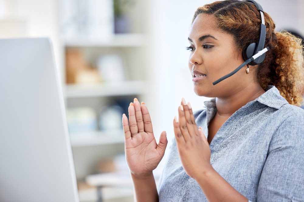 A call center agent wearing headphones and sitting in front of a computer raises their hands on each side while explaining something in pice about CX and EX and great customer experience.